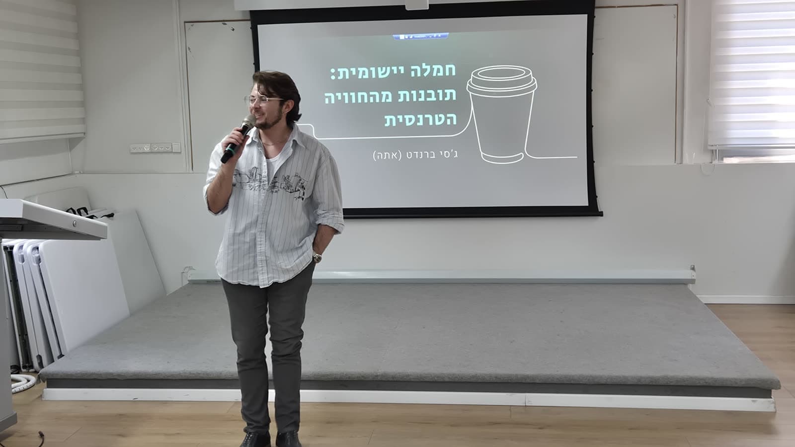 ג'סי ברנדט עומד עם מיקרופון מול מסך שעליו כותרת ההרצאה "חמלה יישומית: תובנות מהחוויה הטרנסית"